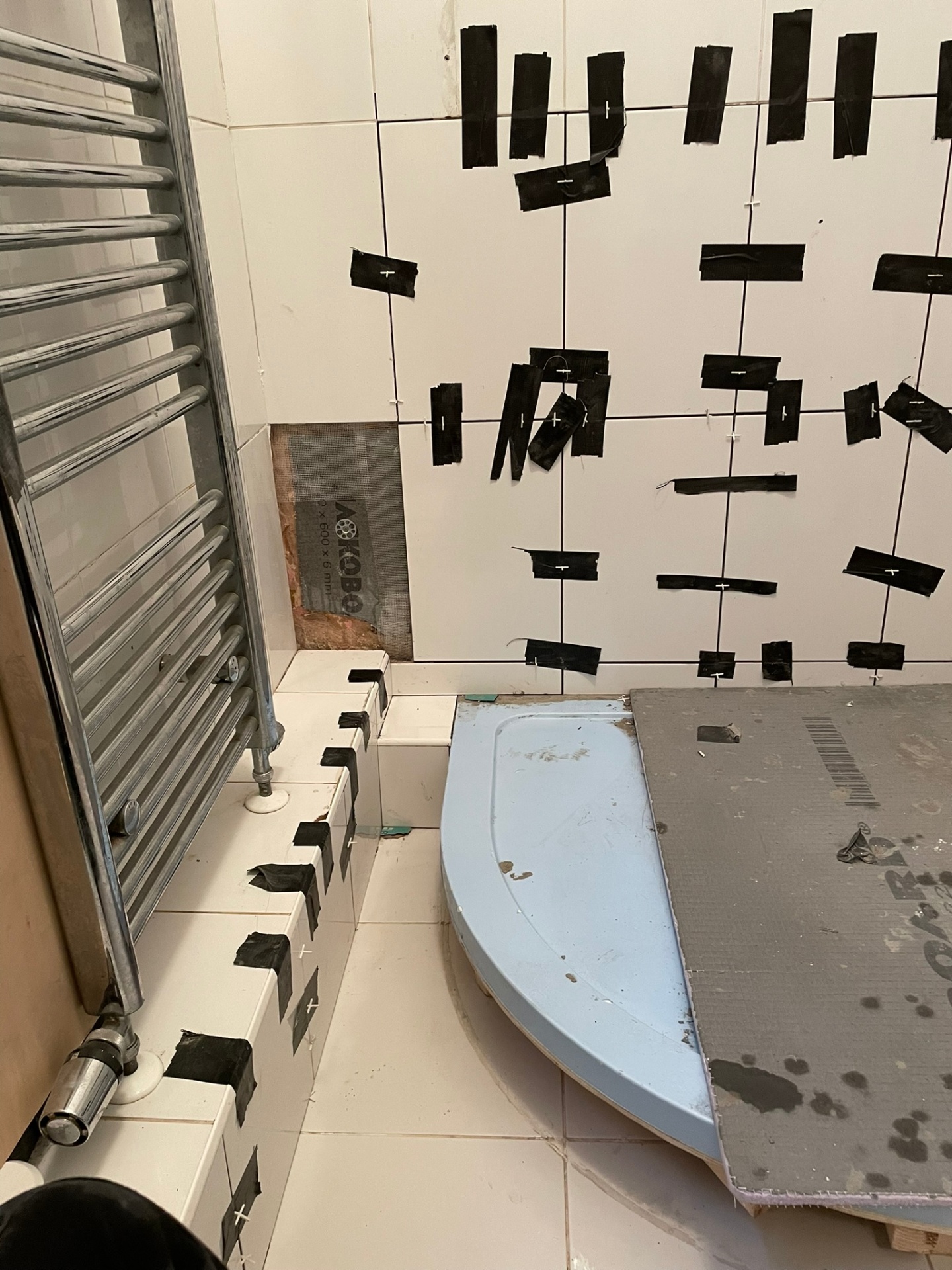 Bathroom tiling in progress showing a blue curved shower tray, towel radiator and tiles held in place with black tape — Ash Villa on-site